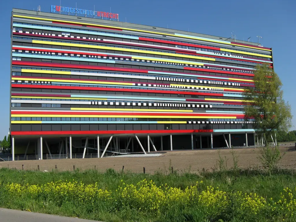 Модерна сграда на Hogeschool Utrecht с цветни хоризонтални ивици по фасадата, зелена трева и жълти цветя отпред